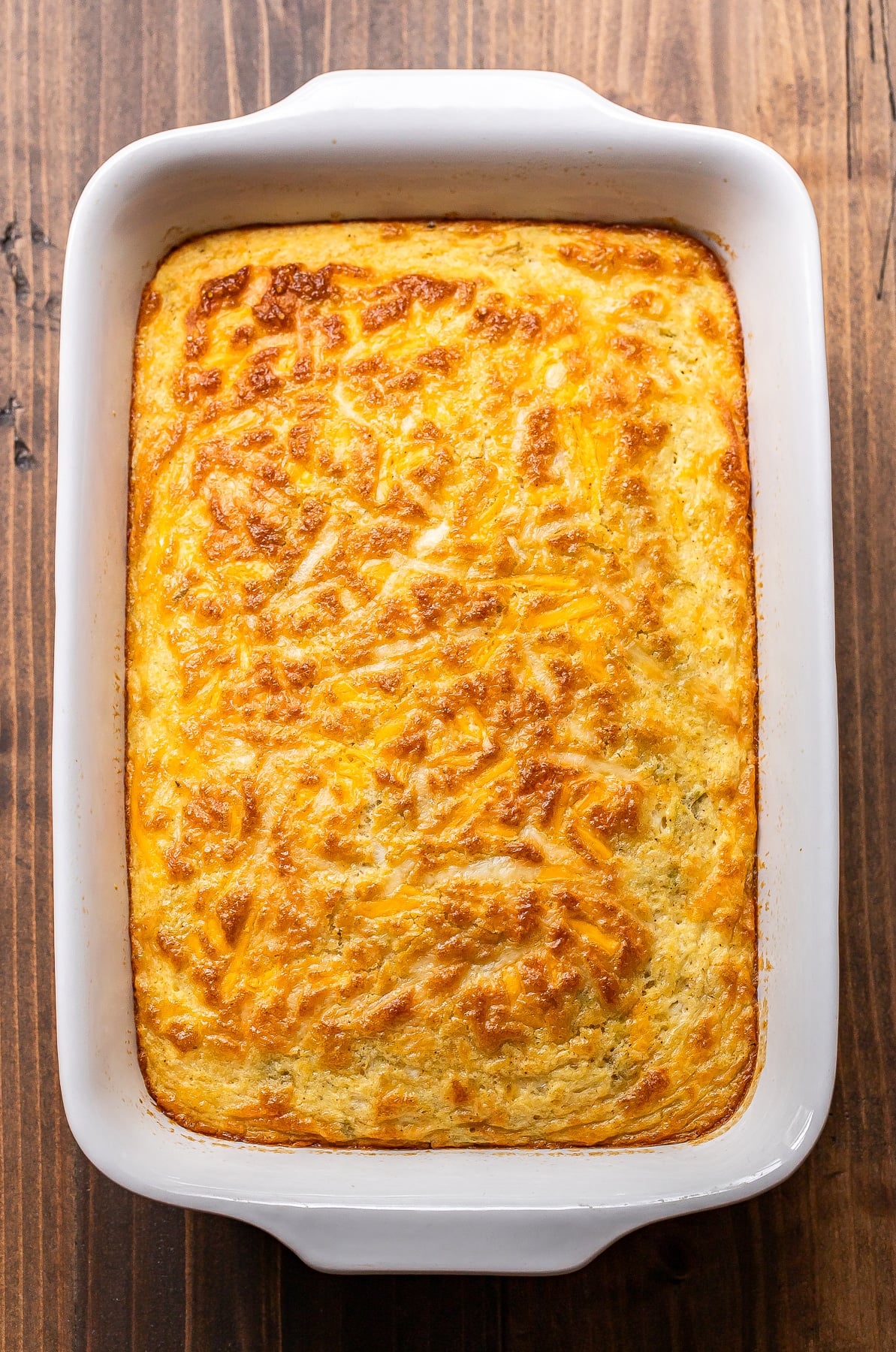 Golden, cheesy cornbread casserole in a white rectangular baking dish, resting on a wooden surface. The top is browned and bubbly, with visible shreds of melted cheese.