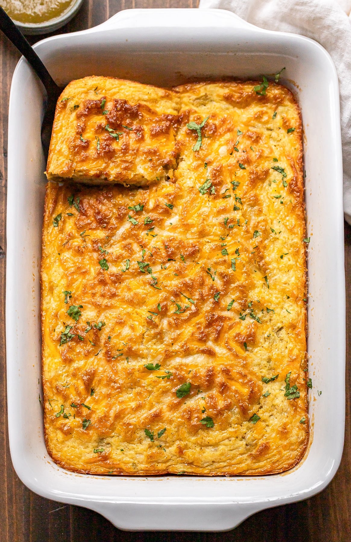 A baked casserole topped with melted cheese and herbs in a white dish, with one square piece cut and a serving spoon next to it.