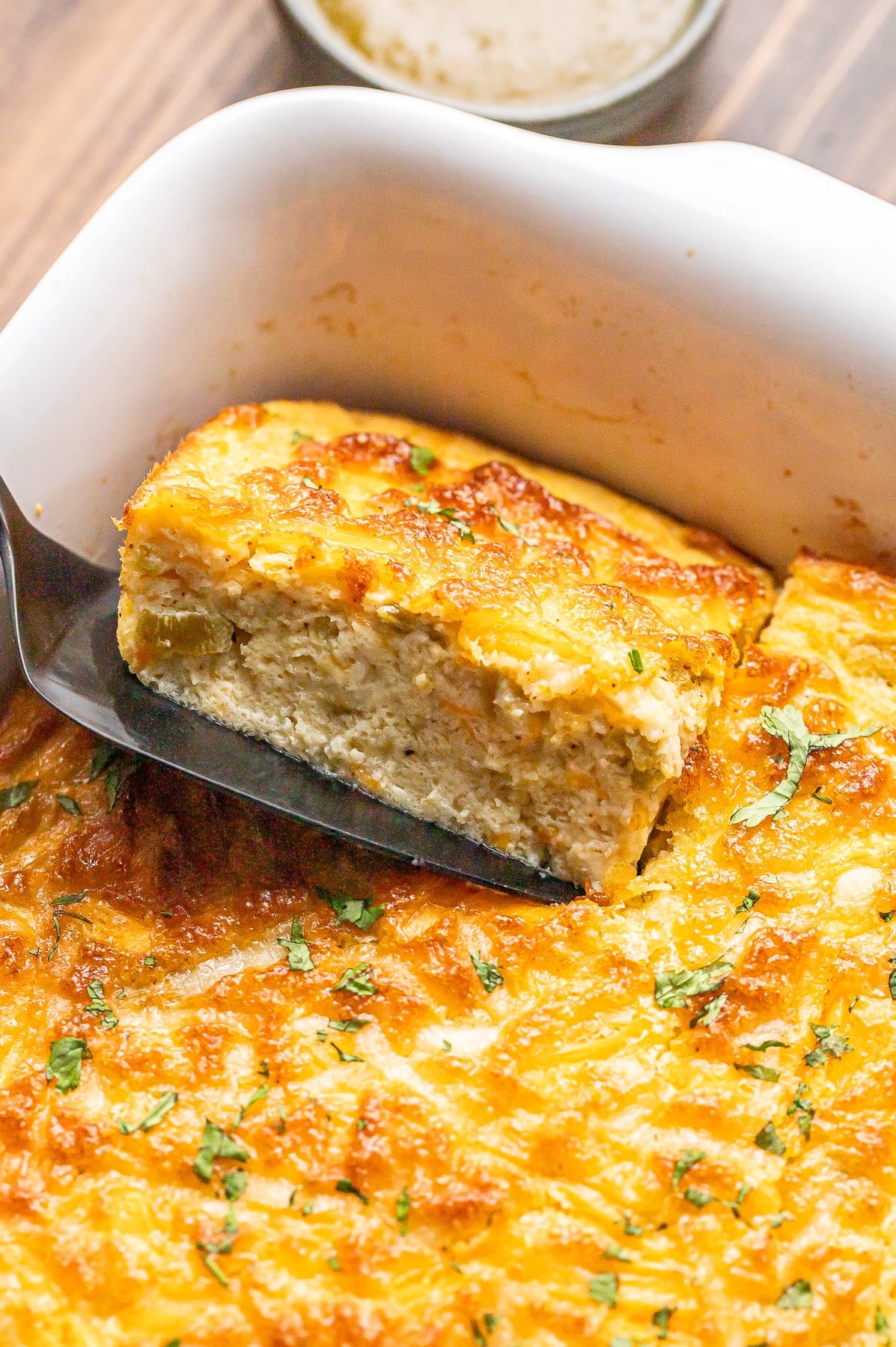 A spatula lifts a cheesy, golden-brown baked casserole from a white dish. The casserole is topped with herbs and appears creamy inside. A small bowl with sauce is visible in the background.