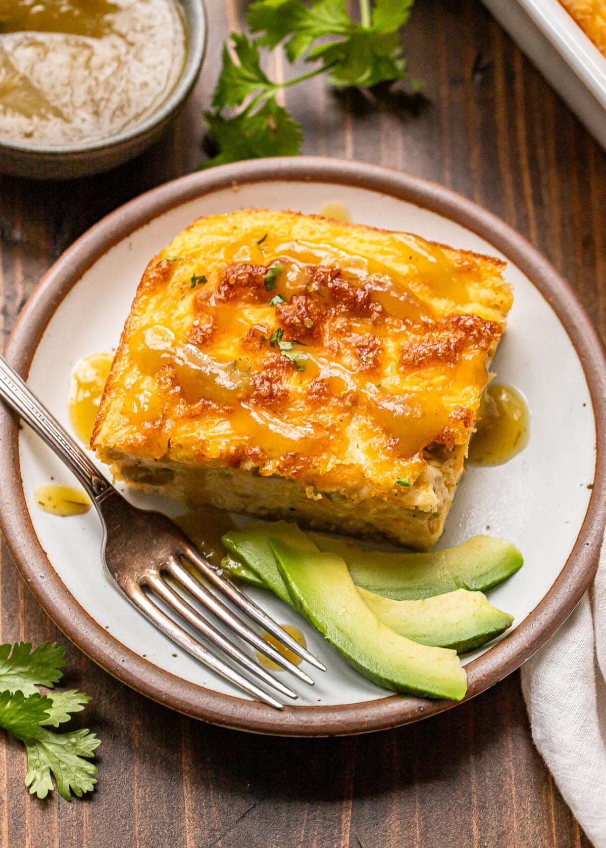 A slice of cheesy casserole topped with sauce sits on a plate beside avocado slices, with a fork and fresh cilantro garnish on a wooden table.