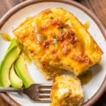 A slice of cheesy breakfast casserole topped with sauce is served on a plate with sliced avocado. A fork holds a bite of the casserole. A small dish of sauce and some parsley are visible in the background.