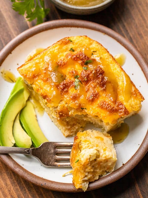 A slice of cheesy breakfast casserole topped with sauce is served on a plate with sliced avocado. A fork holds a bite of the casserole. A small dish of sauce and some parsley are visible in the background.