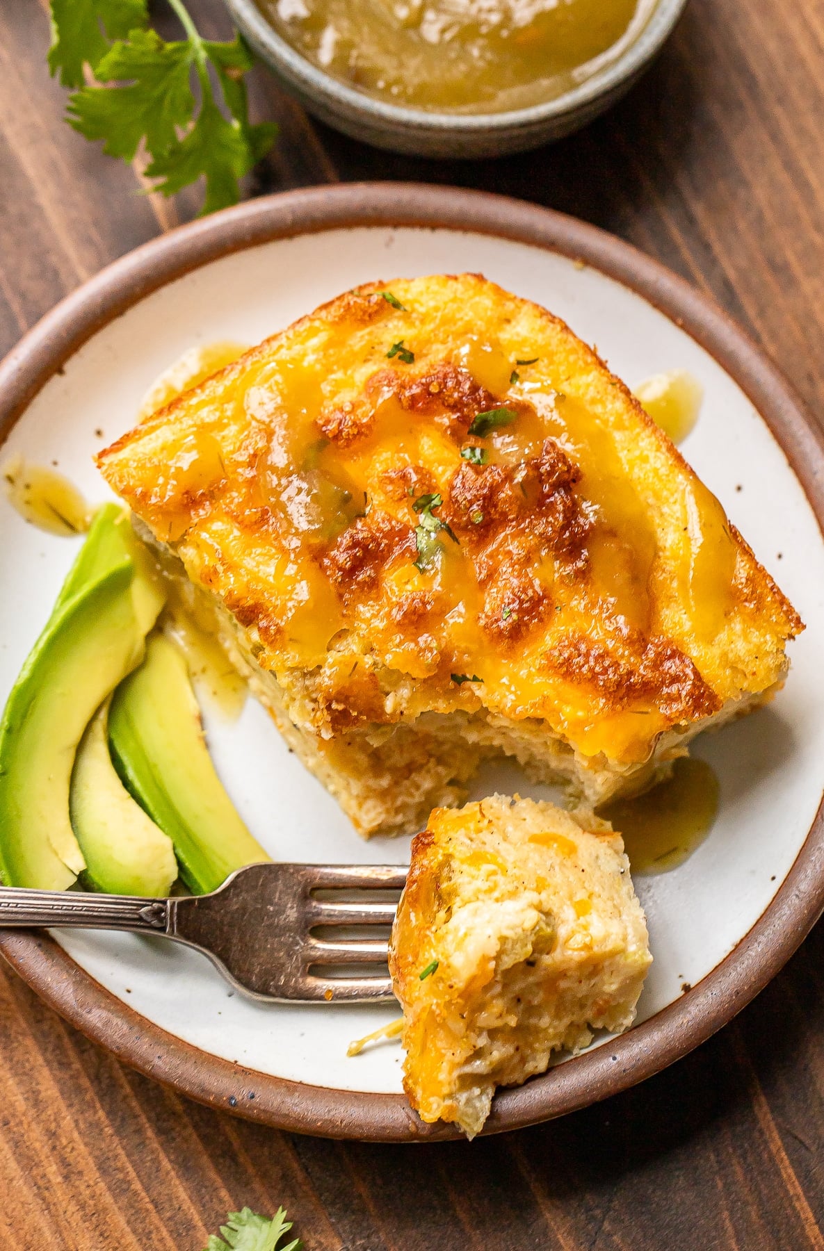 A slice of cheesy breakfast casserole topped with sauce is served on a plate with sliced avocado. A fork holds a bite of the casserole. A small dish of sauce and some parsley are visible in the background.
