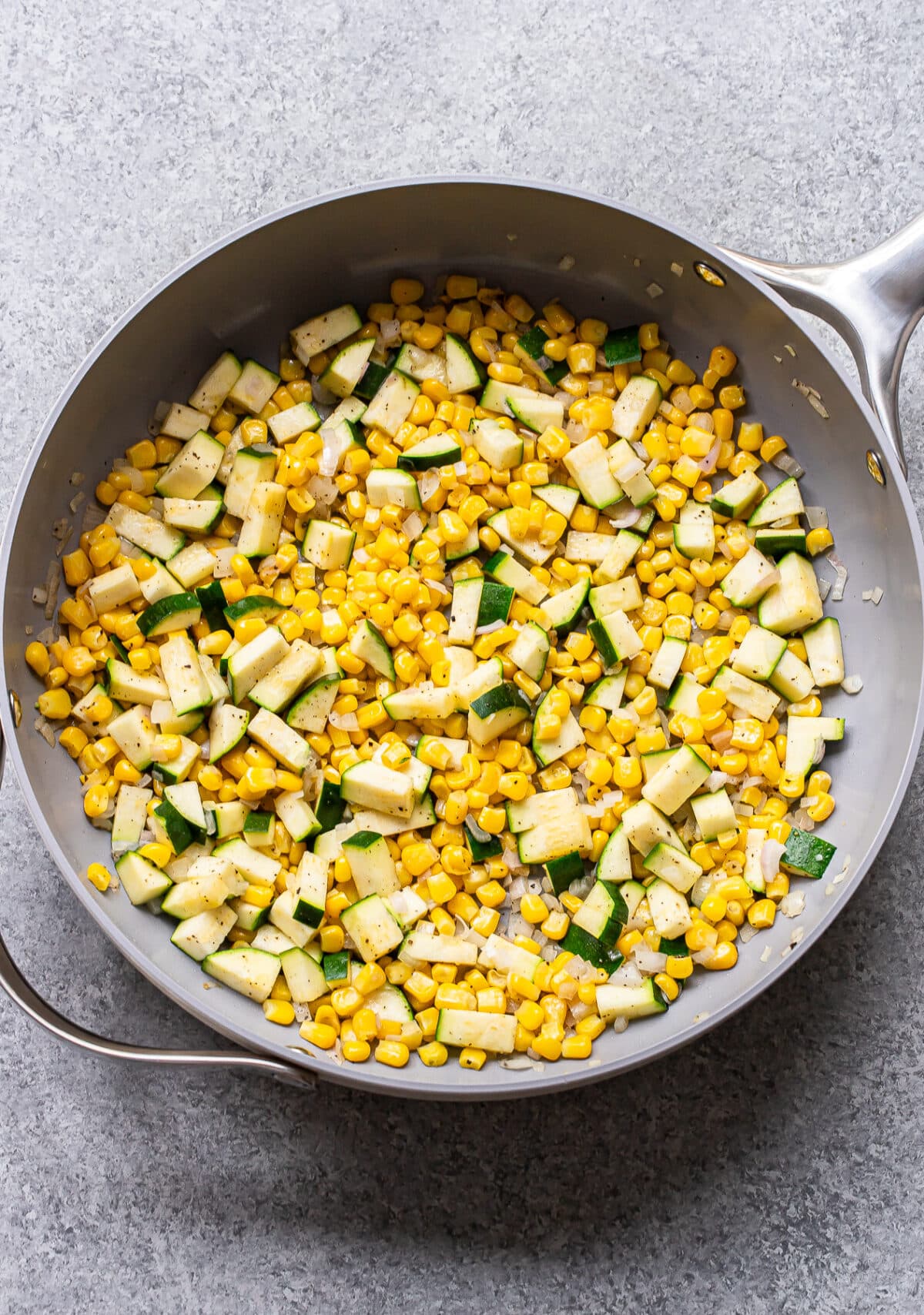 A skillet filled with sautéed corn, diced zucchini, and tender orzo, seasoned with black pepper, on a gray countertop.