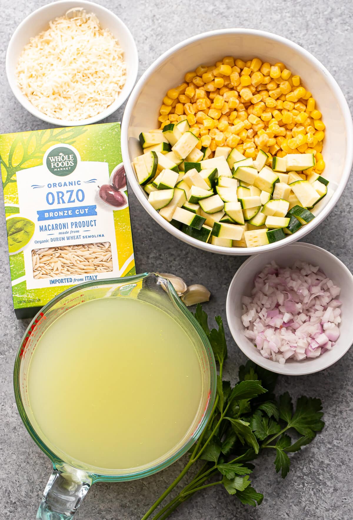Bowls filled with zucchini, corn, diced zucchini, chopped shallot, shredded cheese, a box of Whole Foods orzo pasta, a measuring cup with broth, and fresh parsley are arranged on a gray background—perfect for assembling a flavorful Zucchini Corn Orzo dish.
