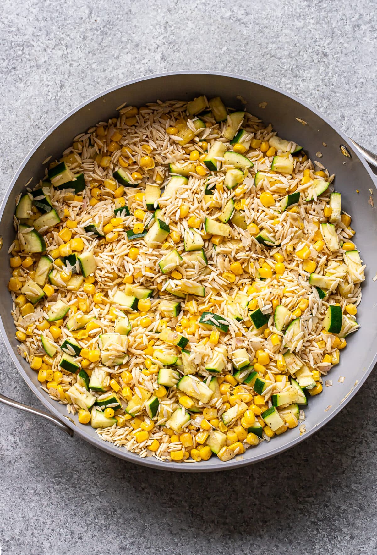 A skillet filled with zucchini corn orzo, artichoke hearts, and tender vegetables, all mixed together on a gray surface.