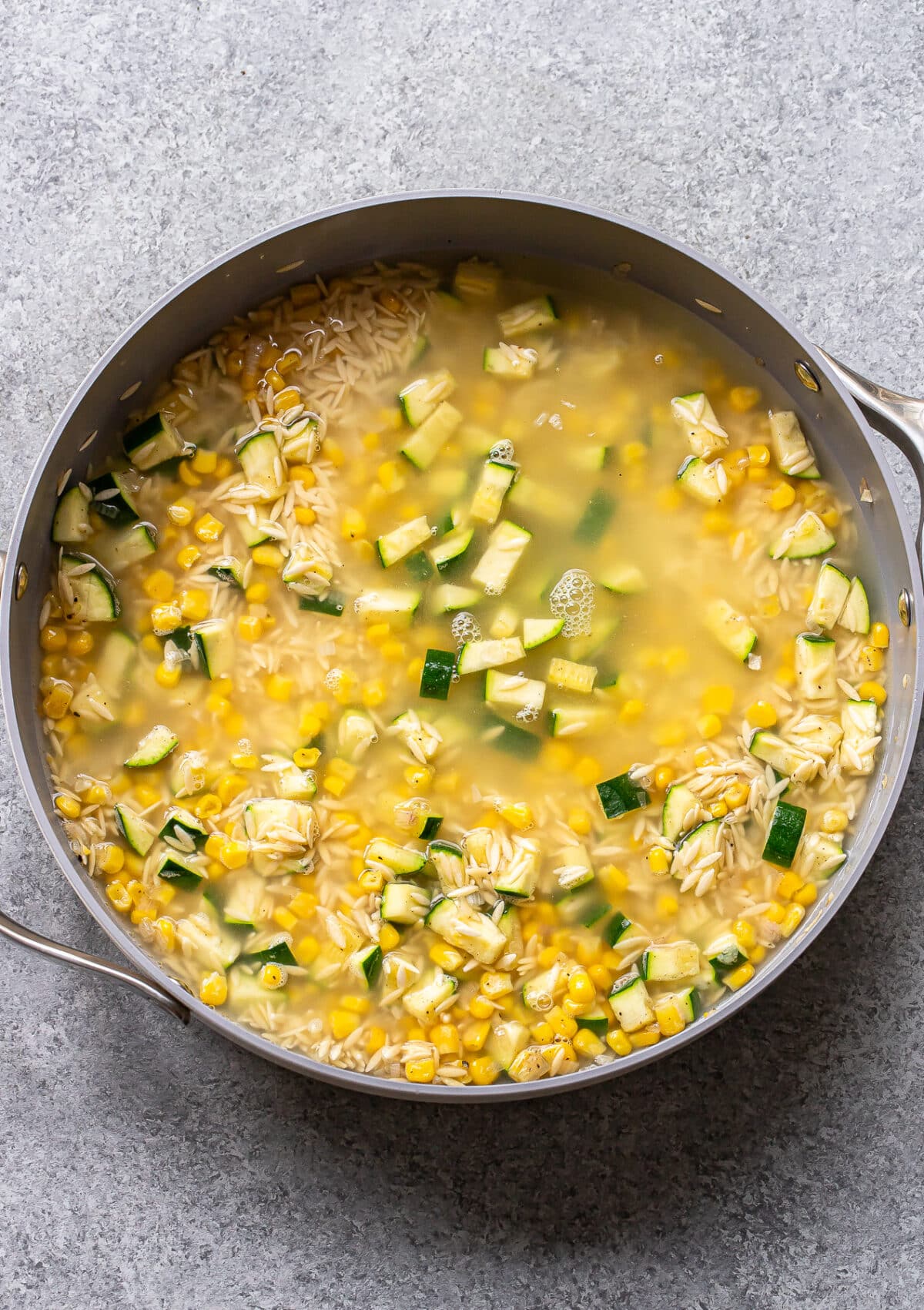 A large pan filled with savory broth, zucchini corn orzo, and tender artichoke hearts sits on a gray countertop.