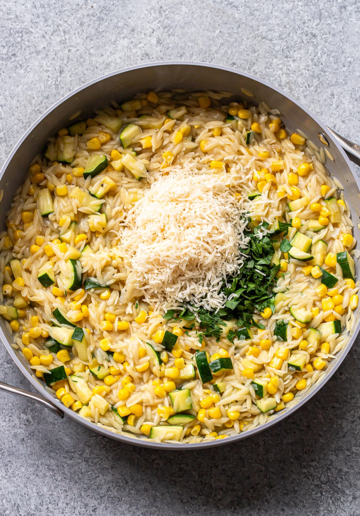 A skillet filled with zucchini corn orzo pasta, topped with shredded cheese and chopped herbs, sits on a gray surface.