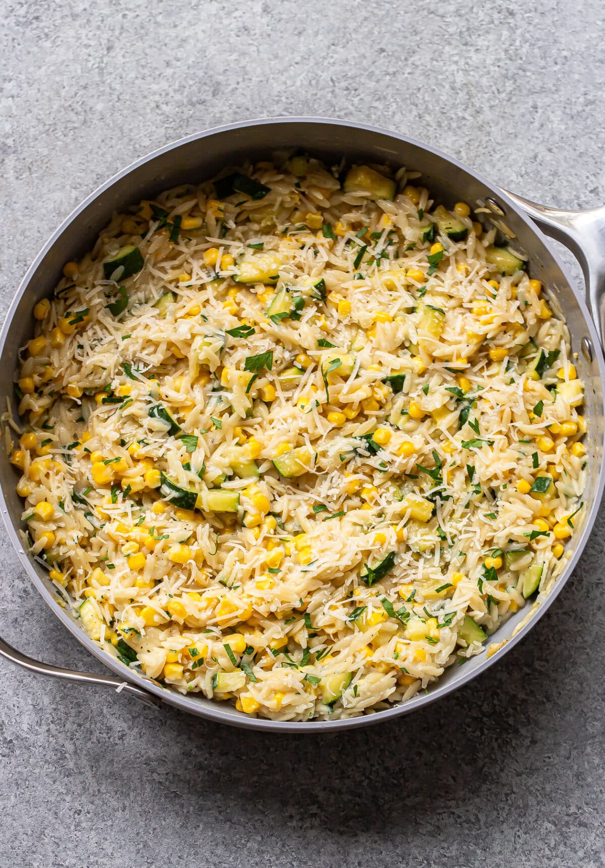 A pan filled with cooked zucchini corn orzo pasta, mixed with fresh herbs and grated cheese, is placed on a gray textured surface.