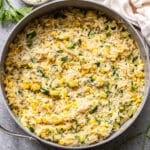 A skillet filled with creamy zucchini corn orzo, mixed with fresh parsley and grated cheese, sits on a gray surface with sprigs of parsley and a bowl of extra cheese nearby.