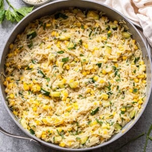 A skillet filled with creamy zucchini corn orzo, mixed with fresh parsley and grated cheese, sits on a gray surface with sprigs of parsley and a bowl of extra cheese nearby.
