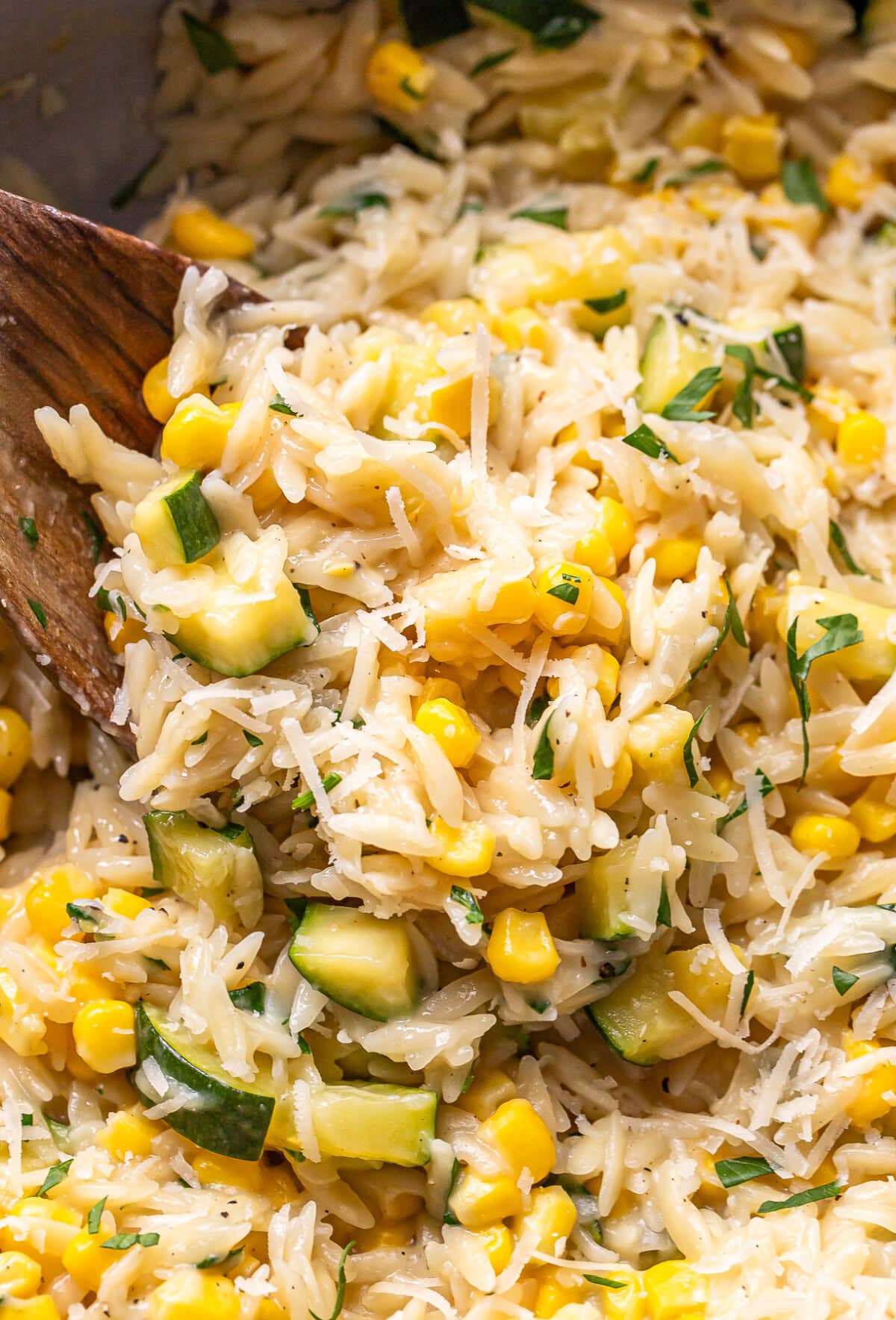 A close-up of a wooden spoon stirring a mixture of rice, zucchini, corn, and fresh herbs. The dish looks creamy and well-seasoned, reminiscent of a savory zucchini corn orzo packed with vibrant flavors.