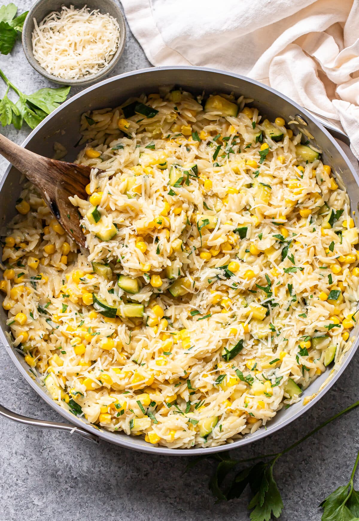 Creamy zucchini corn orzo pasta is mixed with tender corn, sautéed zucchini, fresh herbs, and grated cheese in a large skillet. A wooden spoon is stirring the vibrant mixture. Nearby, a bowl of cheese and fresh parsley await for garnishing this flavorful zucchini corn orzo dish.