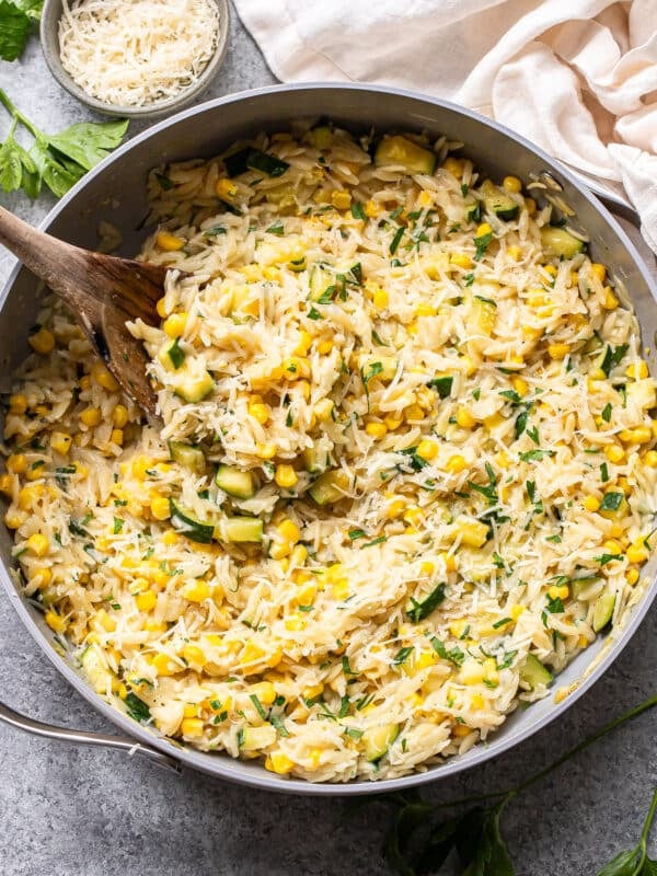 Creamy zucchini corn orzo pasta is mixed with tender corn, sautéed zucchini, fresh herbs, and grated cheese in a large skillet. A wooden spoon is stirring the vibrant mixture. Nearby, a bowl of cheese and fresh parsley await for garnishing this flavorful zucchini corn orzo dish.