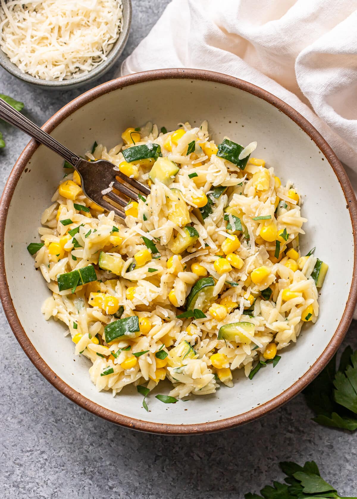 A bowl of Zucchini Corn Orzo pasta salad sits invitingly, featuring tender orzo tossed with sweet corn, chopped zucchini, fresh parsley, and a sprinkle of grated cheese. A fork rests inside the bowl, while a small dish of extra cheese and a neatly folded white napkin are nearby on the gray surface.