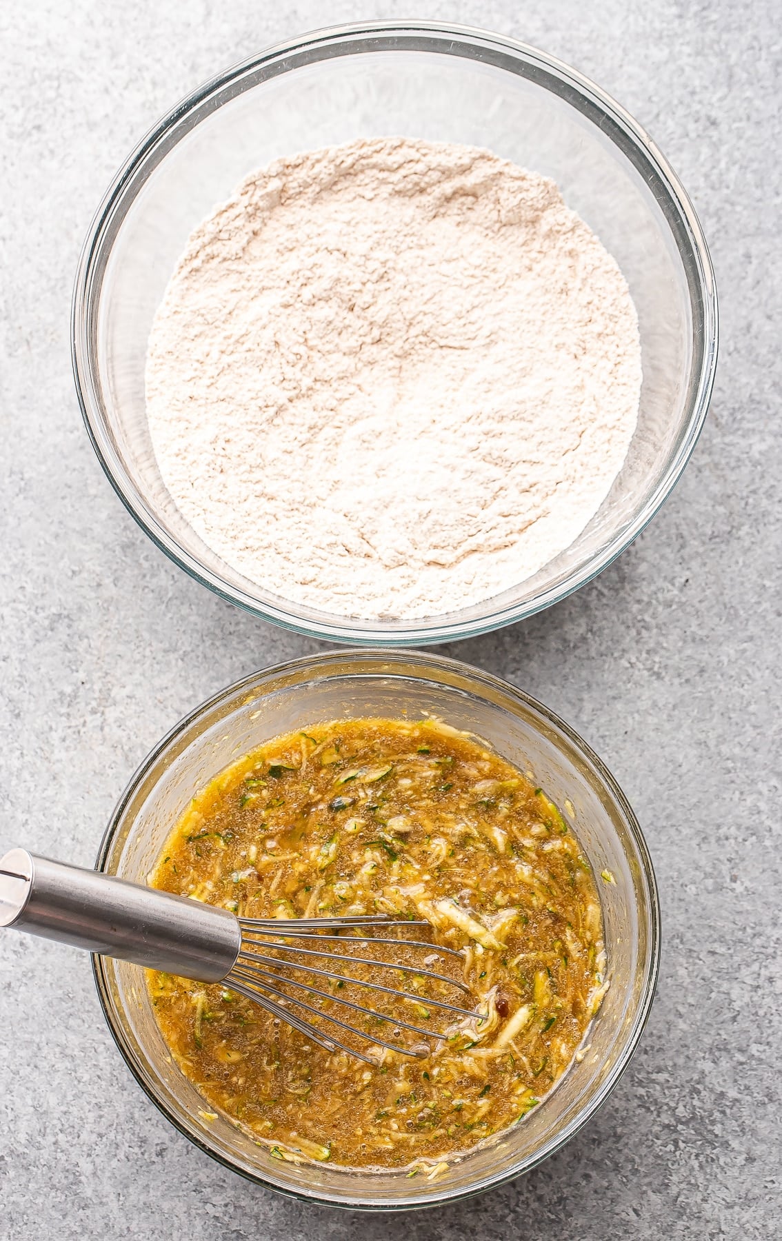 Two glass bowls on a gray surface: one filled with dry flour mixture, the other with a wet zucchini muffin batter being whisked with a metal whisk.