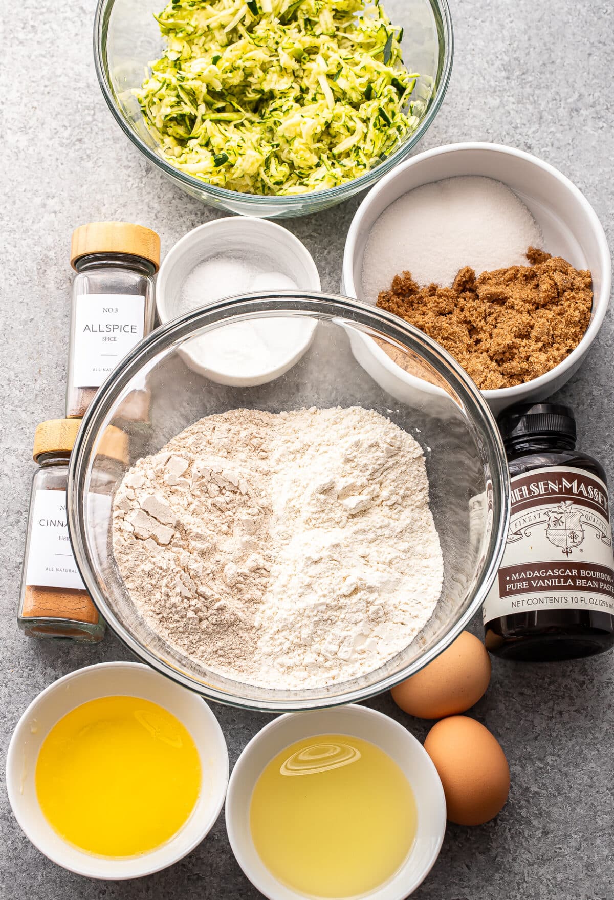Baking ingredients on a countertop for Zucchini Muffins: flour, shredded zucchini, brown and white sugar, eggs, oil, melted butter, vanilla extract, cinnamon, allspice, and baking powder—each measured out in bowls or jars.