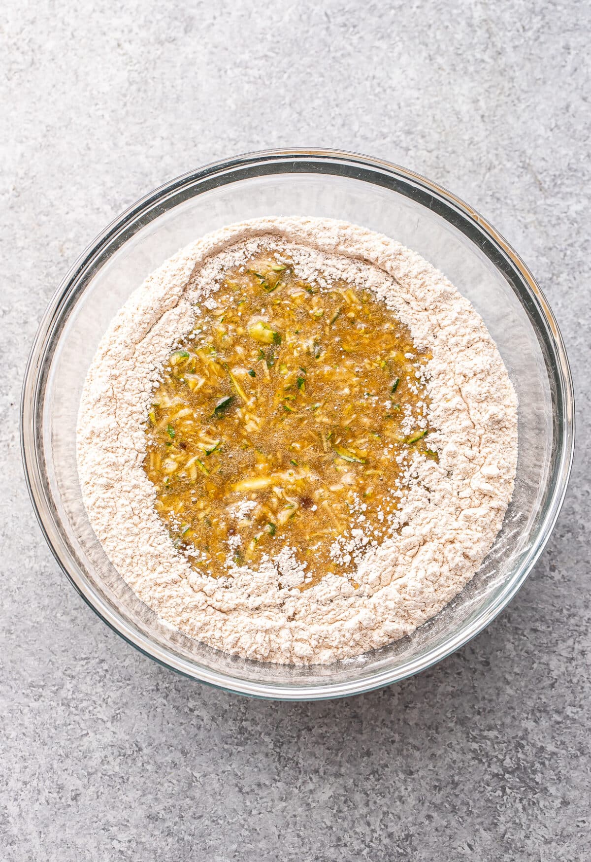 A glass bowl with flour surrounding a mixture of shredded zucchini, eggs, and other wet ingredients—ready to be combined for baking delicious Zucchini Muffins—sits on a gray countertop.