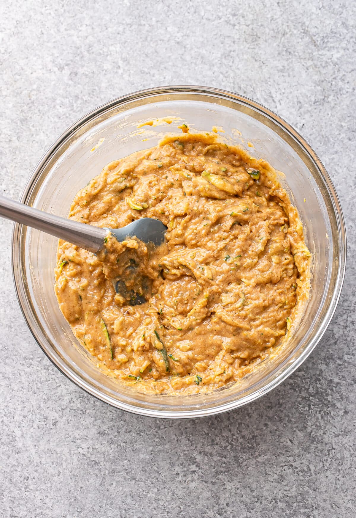A glass bowl filled with thick, light brown zucchini muffin batter and a gray spatula sitting inside, all placed on a light gray textured surface.