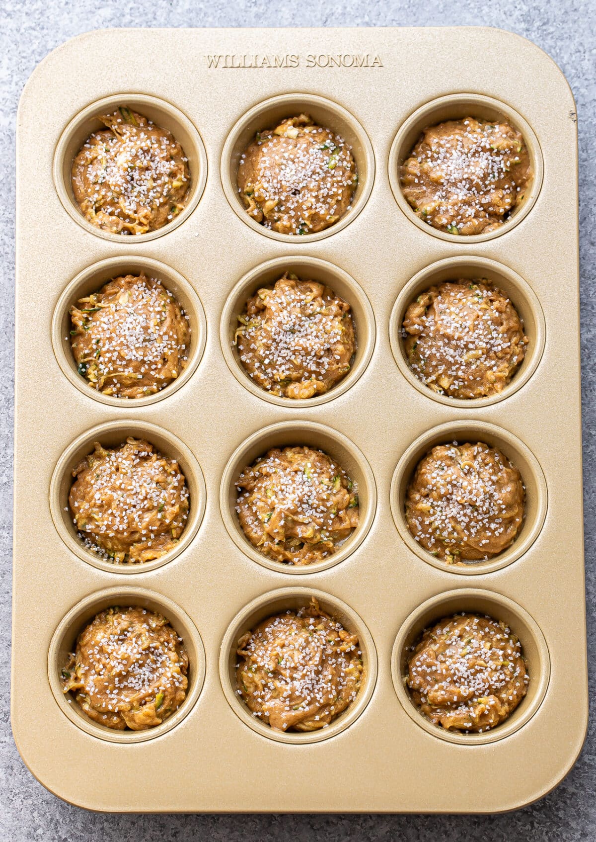 A gold muffin tin holds twelve unbaked Zucchini Muffins batter portions, each topped with a dusting of granulated sugar. The batter appears to have visible zucchini shreds. The muffin tin is embossed with Williams Sonoma.