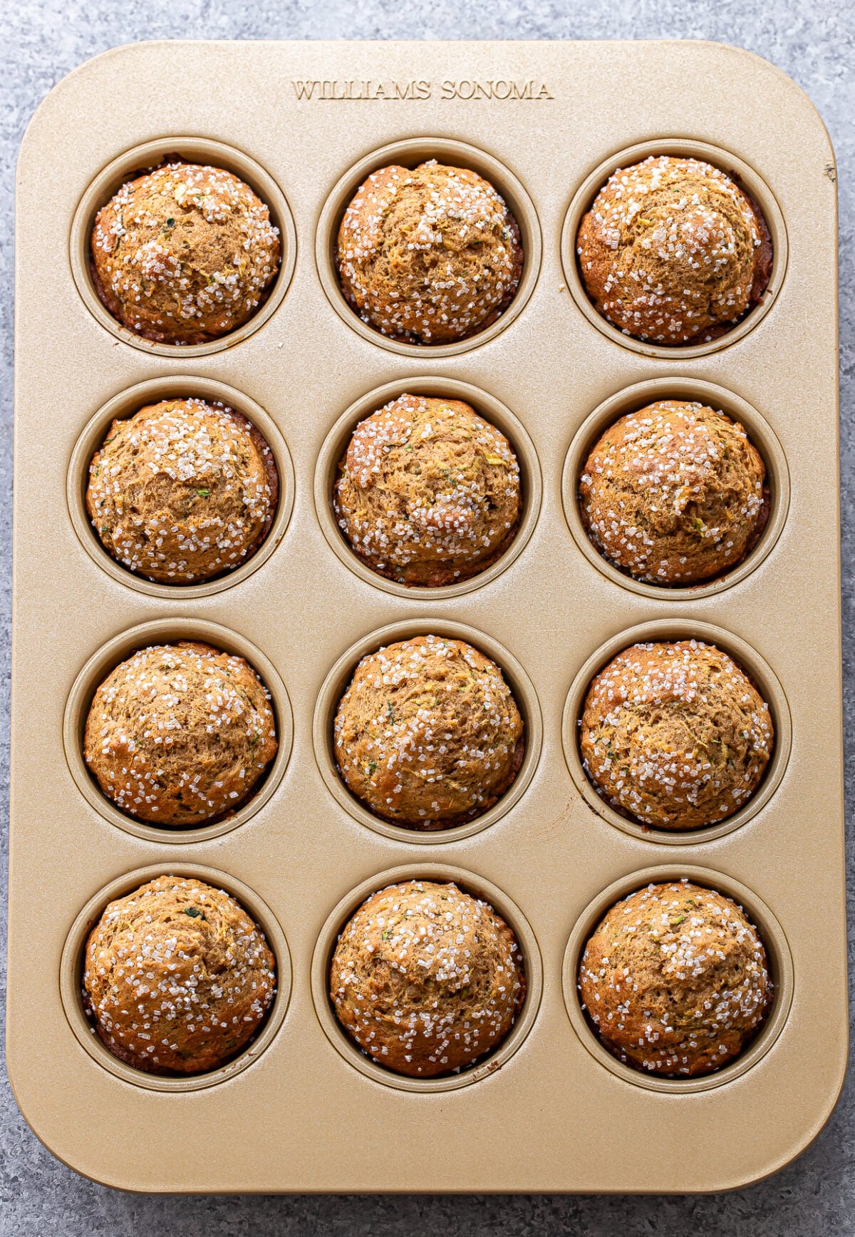 A muffin tin with 12 golden-brown zucchini muffins topped with coarse sugar sits on a gray countertop. The tin is marked WILLIAMS SONOMA at the top.