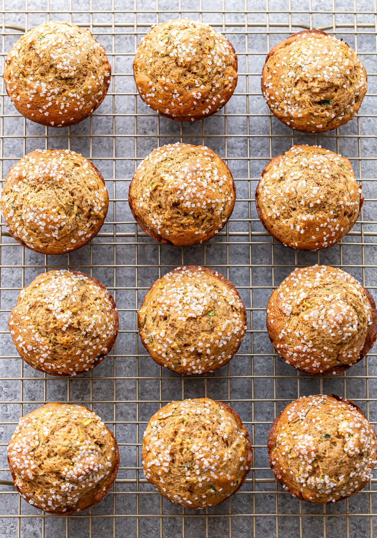 Twelve golden brown zucchini muffins topped with coarse sugar are cooling on a metal wire rack, arranged in neat rows on a gray countertop.