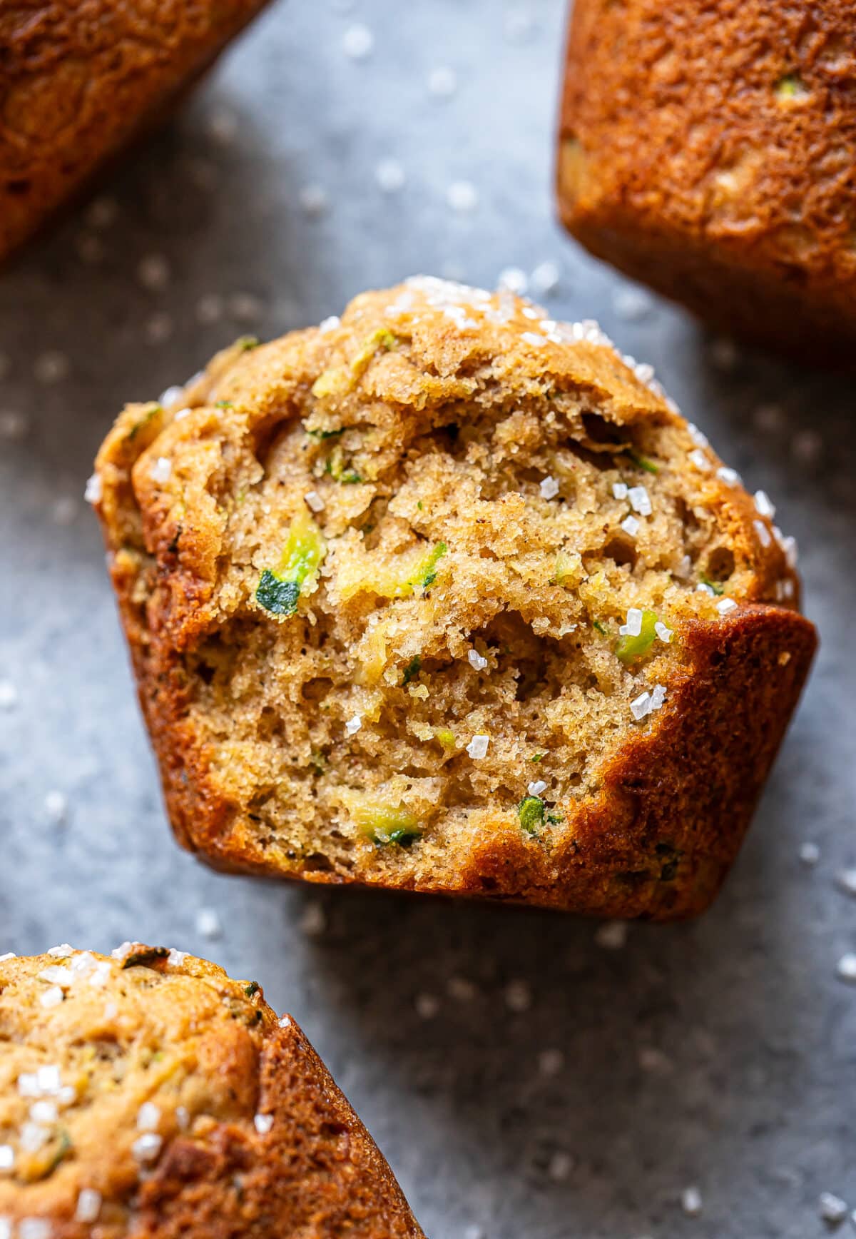 Close-up of a golden-brown Zucchini Muffin topped with coarse sugar, showing a moist, textured crumb and visible green zucchini pieces, on a gray surface with other zucchini muffins partially visible nearby.