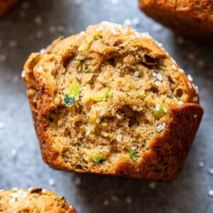 Close-up of a golden-brown Zucchini Muffin topped with coarse sugar, showing a moist, textured crumb and visible green zucchini pieces, on a gray surface with other zucchini muffins partially visible nearby.