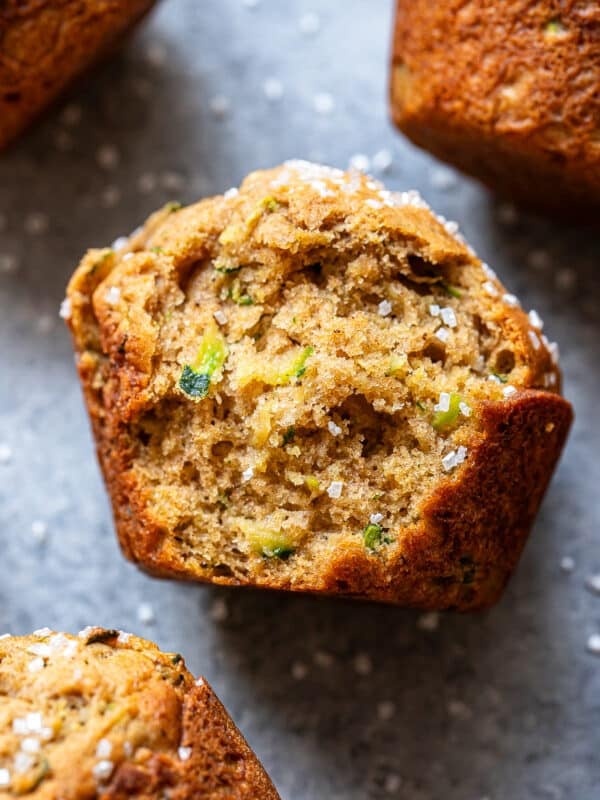Close-up of a golden-brown Zucchini Muffin topped with coarse sugar, showing a moist, textured crumb and visible green zucchini pieces, on a gray surface with other zucchini muffins partially visible nearby.