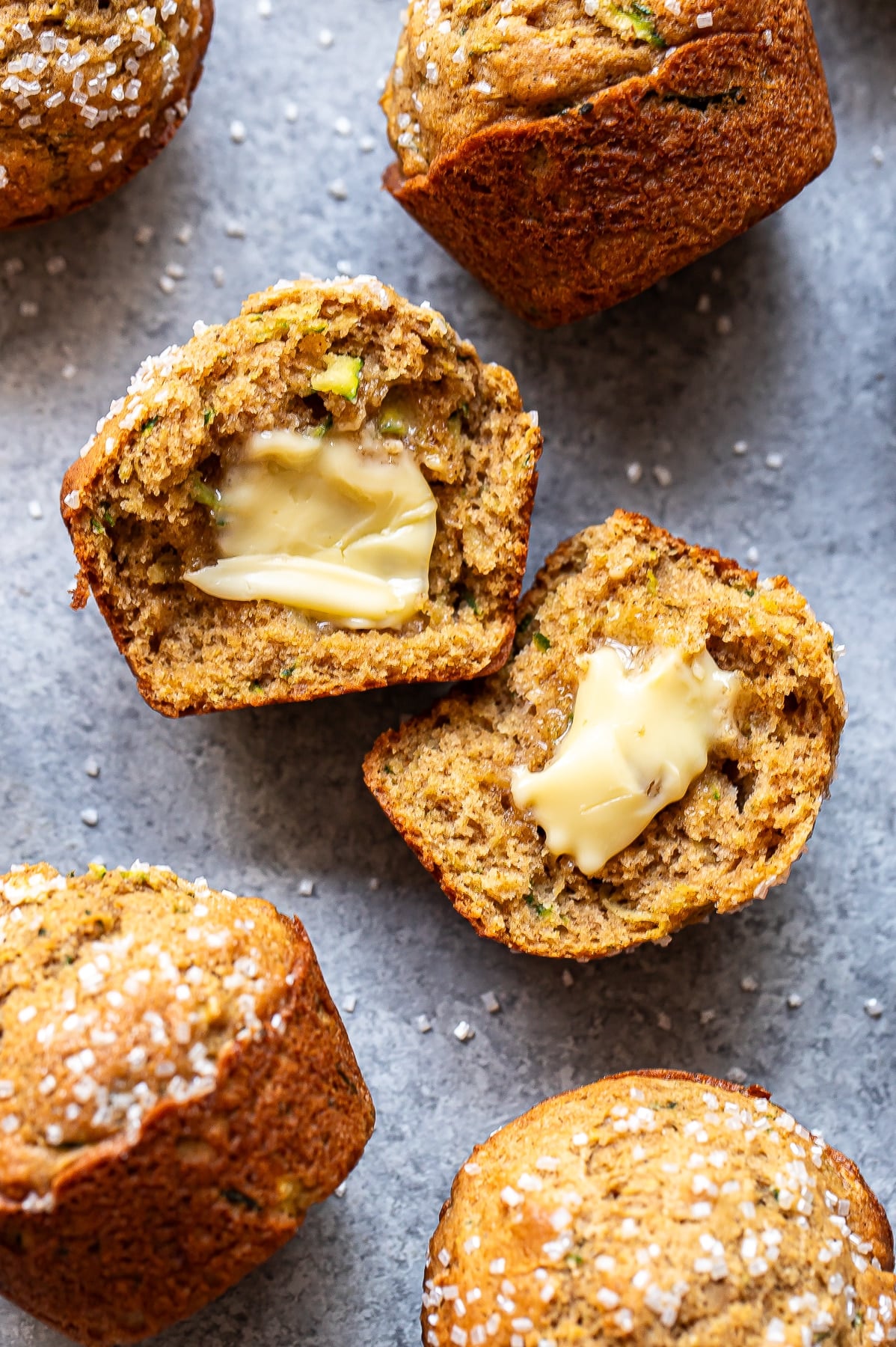 Four Zucchini Muffins rest on a gray surface, two split open to reveal melted butter inside. The muffins are topped with coarse sugar and have a moist, textured interior that highlights the fresh flavor of zucchini.