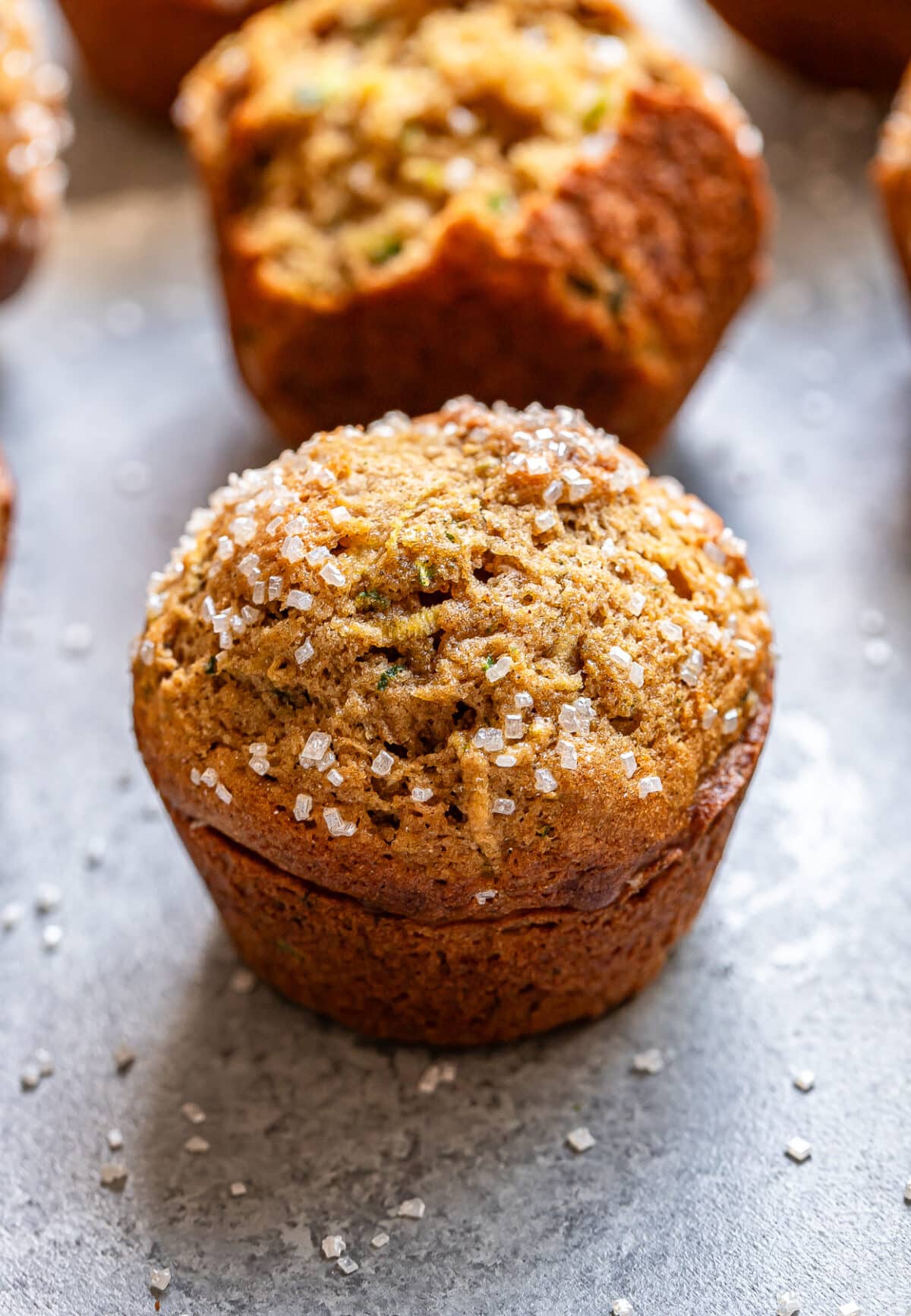 A close-up of a golden brown zucchini muffin sprinkled with coarse sugar on top, sitting on a gray surface with more zucchini muffins blurred in the background.