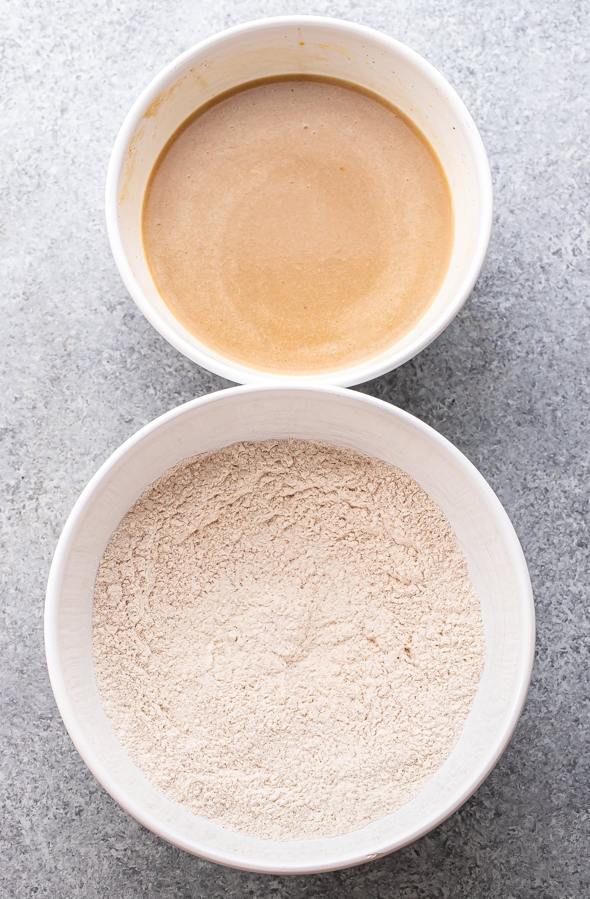 Two white bowls on a gray surface: the top bowl contains a creamy liquid mixture, while the bottom bowl holds a mound of dry flour mixture, ready to be combined for delicious Apple Cinnamon Muffins.