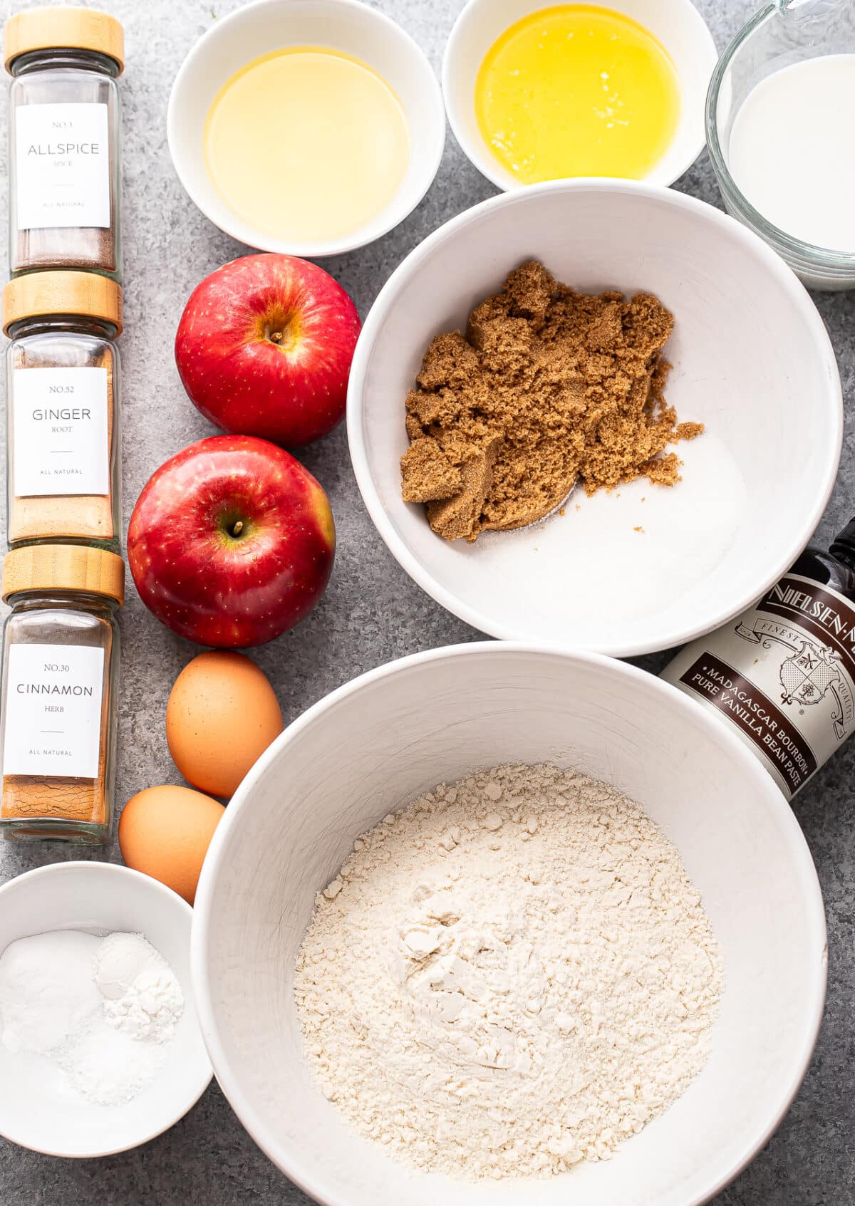 Baking ingredients for Apple Cinnamon Muffins—including apples, flour, brown sugar, eggs, melted butter, vanilla extract, milk, oil, spices (allspice, ginger, cinnamon), baking powder, baking soda, and salt—arranged on a countertop.