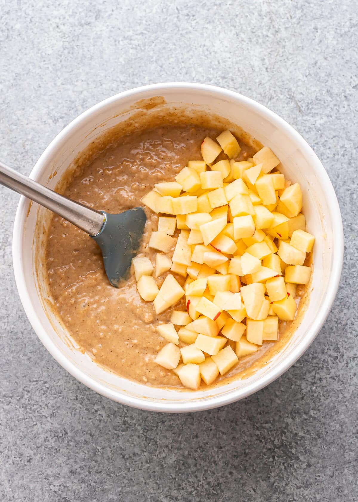 A mixing bowl with Apple Cinnamon Muffins batter and chopped apple pieces on top, being stirred with a spatula on a gray countertop.