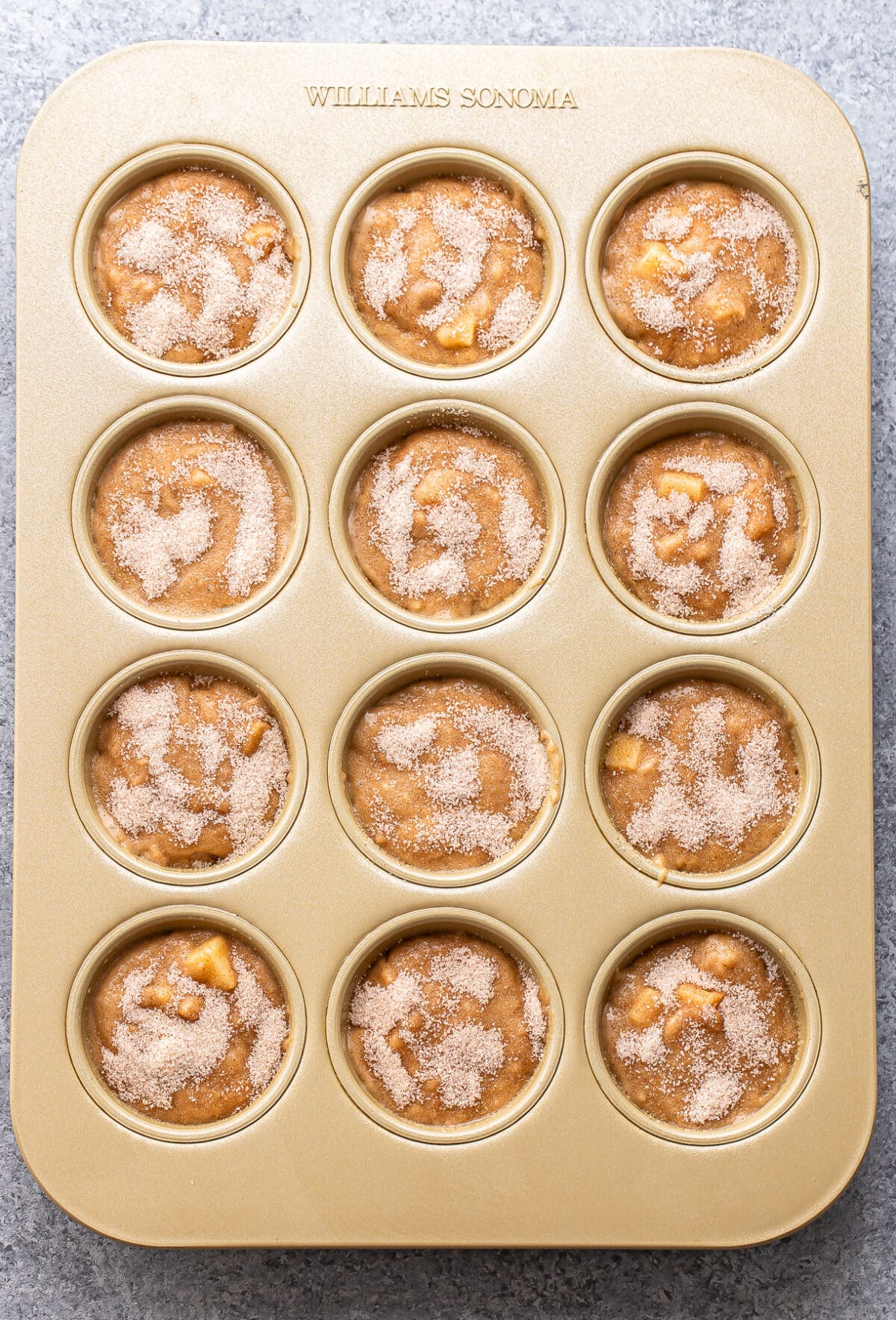A gold muffin tin holds twelve portions of unbaked Apple Cinnamon Muffin batter, each sprinkled with cinnamon sugar on top, ready to be baked. The tin is placed on a gray surface.