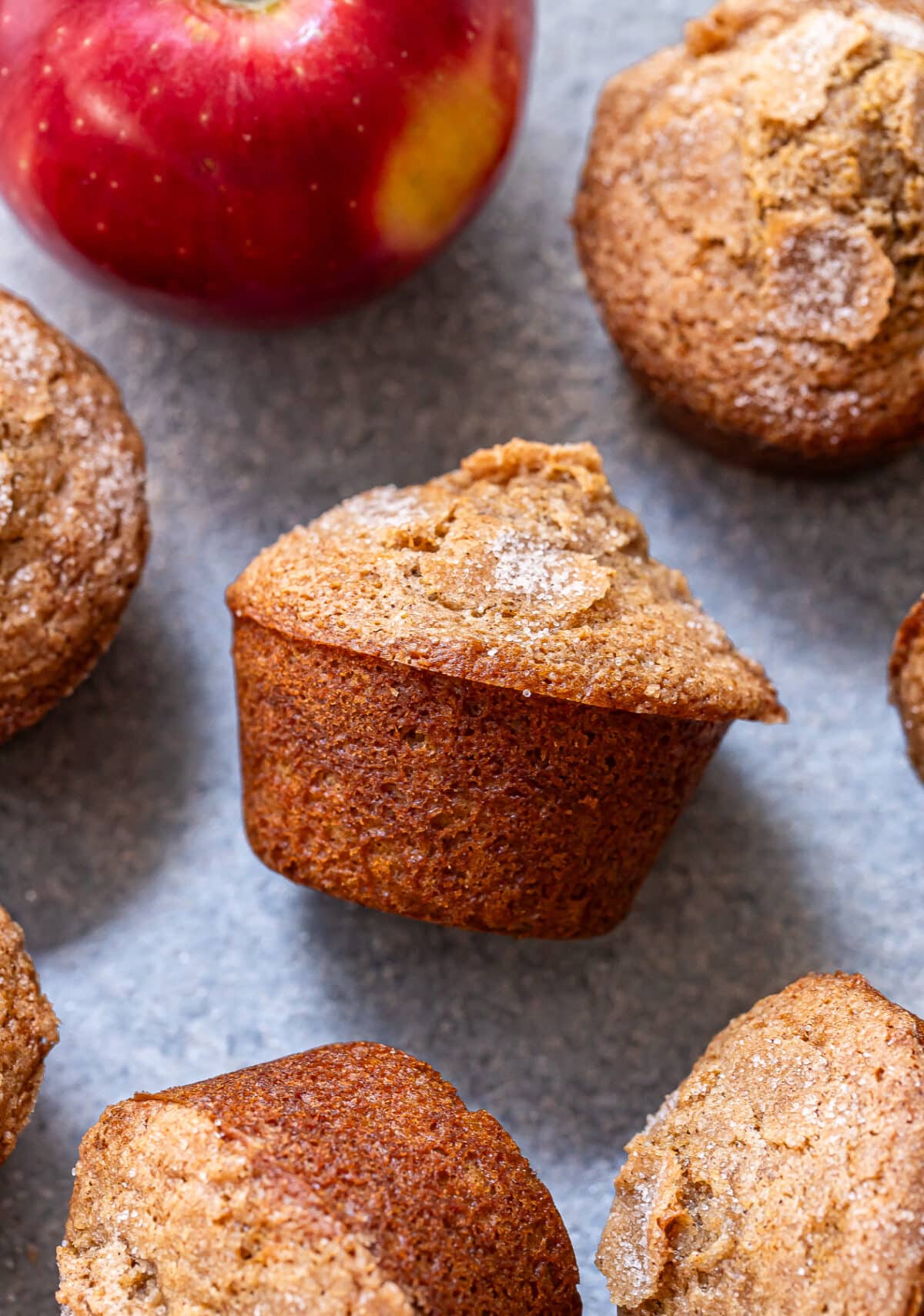 Apple Cinnamon Muffins with a sugared top are arranged on a gray surface next to a red apple.