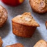 Apple Cinnamon Muffins with a sugared top are arranged on a gray surface next to a red apple.