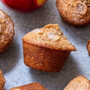 Apple Cinnamon Muffins with a sugared top are arranged on a gray surface next to a red apple.