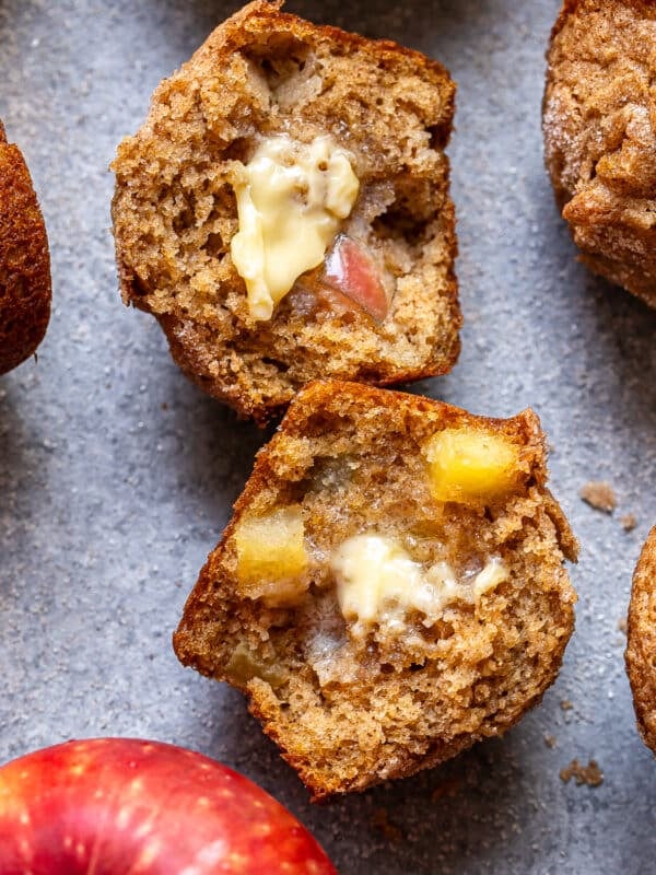 A sliced Apple Cinnamon Muffin with melted butter inside, showing chunks of apple. Surrounding are whole muffins and a fresh red apple on a gray surface.