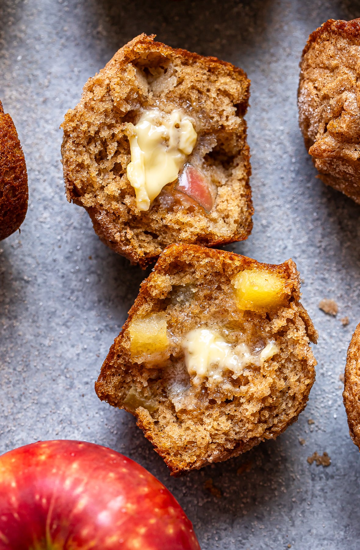 A sliced Apple Cinnamon Muffin with melted butter inside, showing chunks of apple. Surrounding are whole muffins and a fresh red apple on a gray surface.