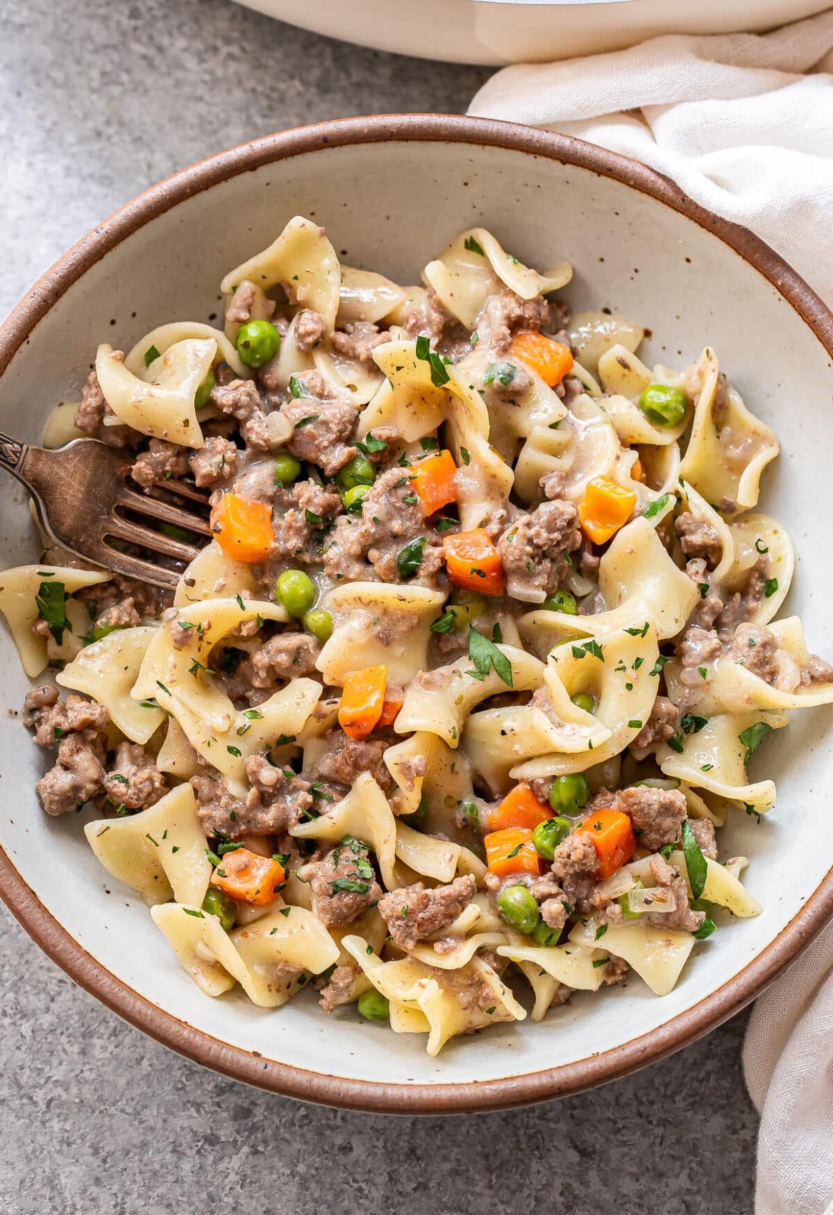 A ceramic bowl filled with egg noodles, ground beef, peas, and diced carrots in a creamy sauce, garnished with chopped herbs. A fork rests inside the bowl.