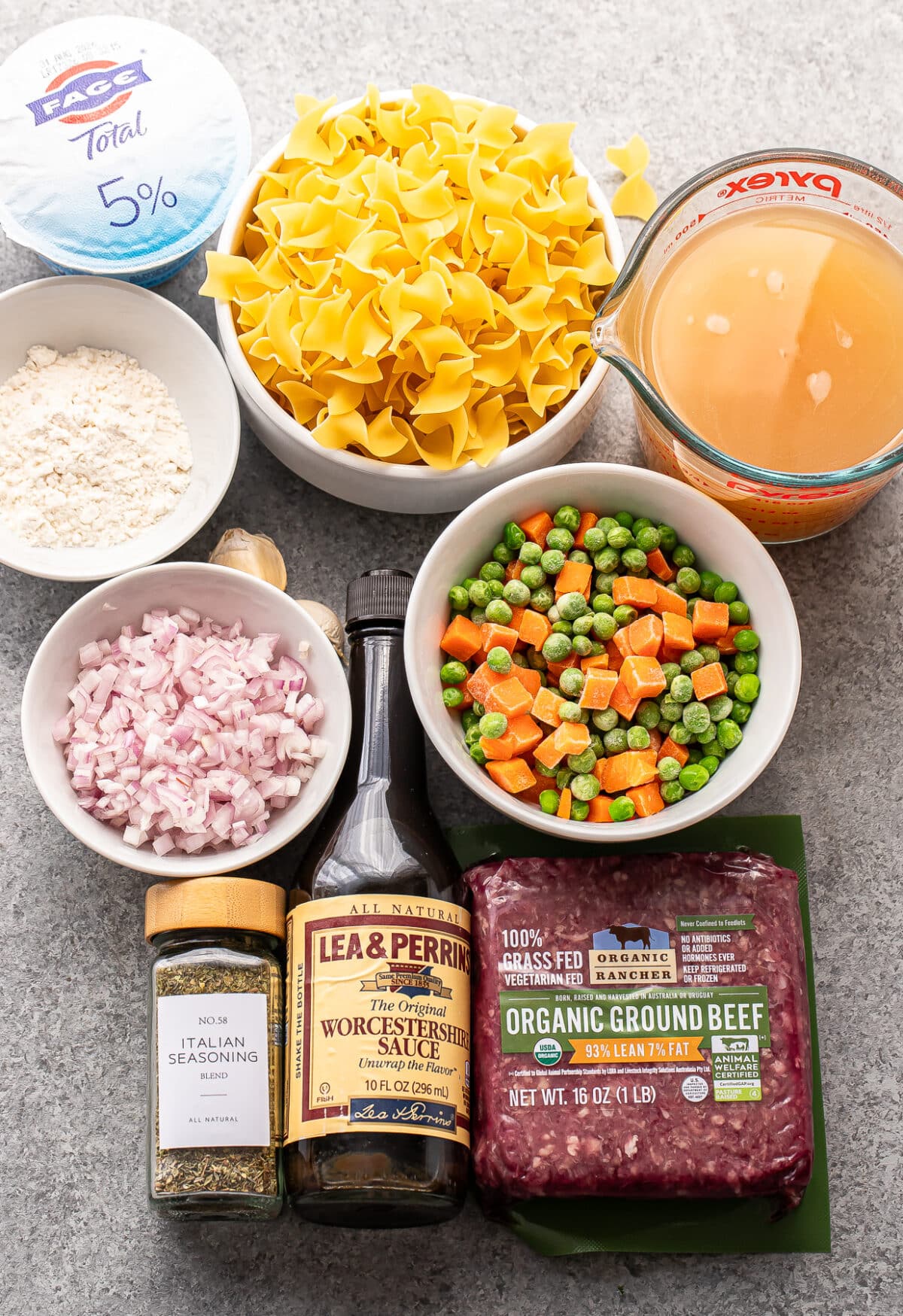 A top-down view of ingredients for a beef noodle recipe: ground beef, egg noodles, frozen mixed vegetables, beef broth, chopped shallots, flour, Italian seasoning, Worcestershire sauce, and Greek yogurt.
