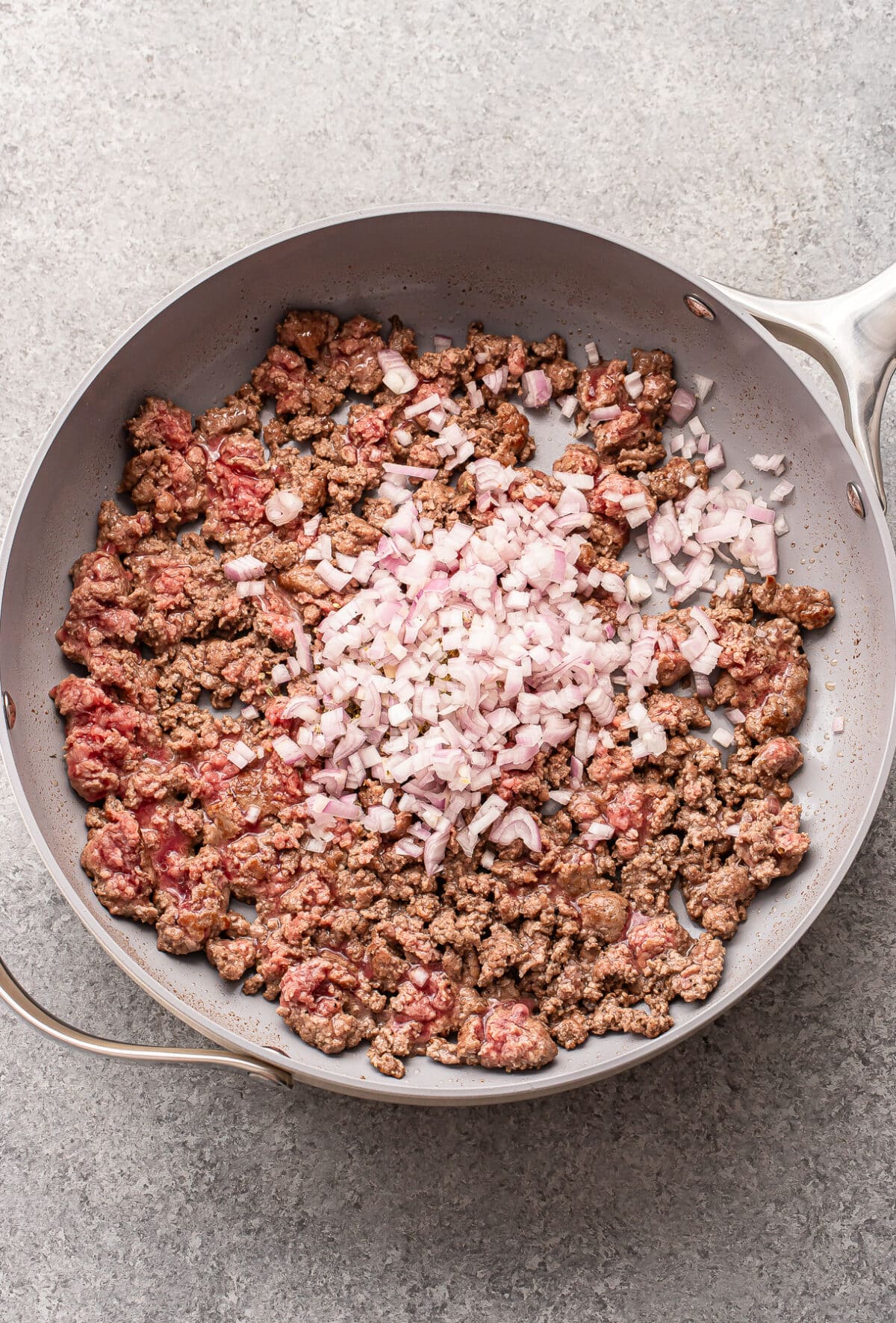 Ground beef is browning in a large skillet, with a pile of chopped onions added in the center, ready to be mixed in. The pan sits on a light-colored countertop.