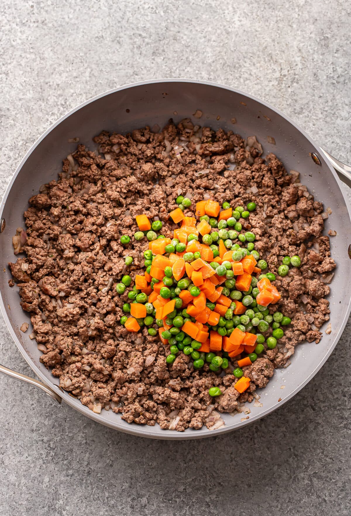 A skillet with cooked ground beef, topped with a pile of diced carrots and green peas, on a gray surface.