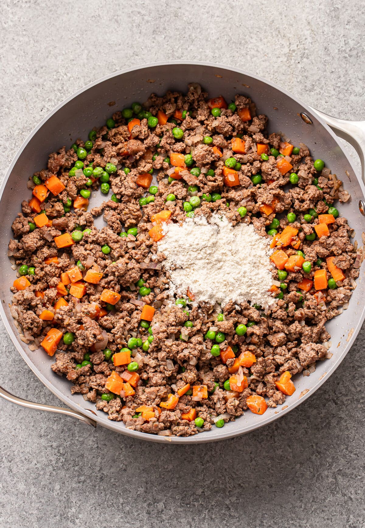 A skillet with cooked ground beef, diced carrots, green peas, and a pile of flour in the center, all on a gray countertop.
