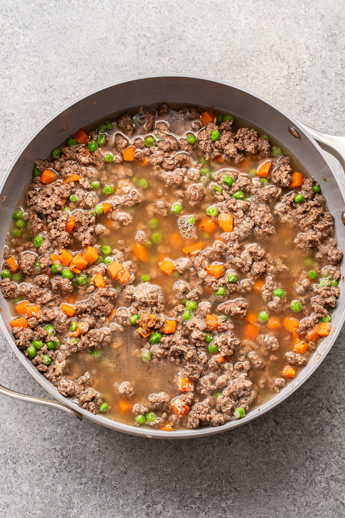 A skillet filled with ground beef, diced carrots, and green peas simmering in a brown broth on a gray surface.