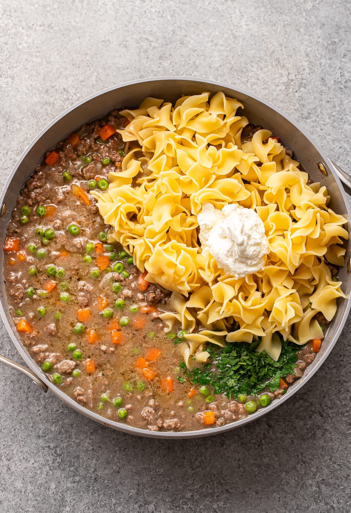 A skillet with cooked ground beef, peas, carrots, uncooked egg noodles, a dollop of sour cream, and chopped parsley, ready to be mixed together.