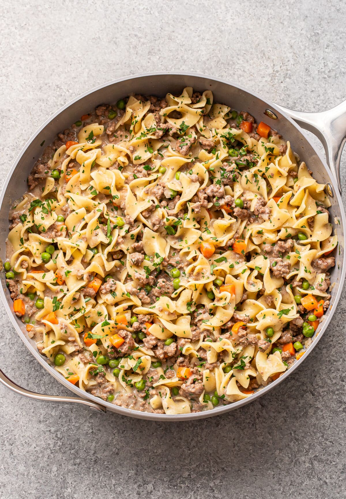 A large skillet filled with creamy beef and noodle casserole, mixed with peas, carrots, and garnished with chopped parsley.