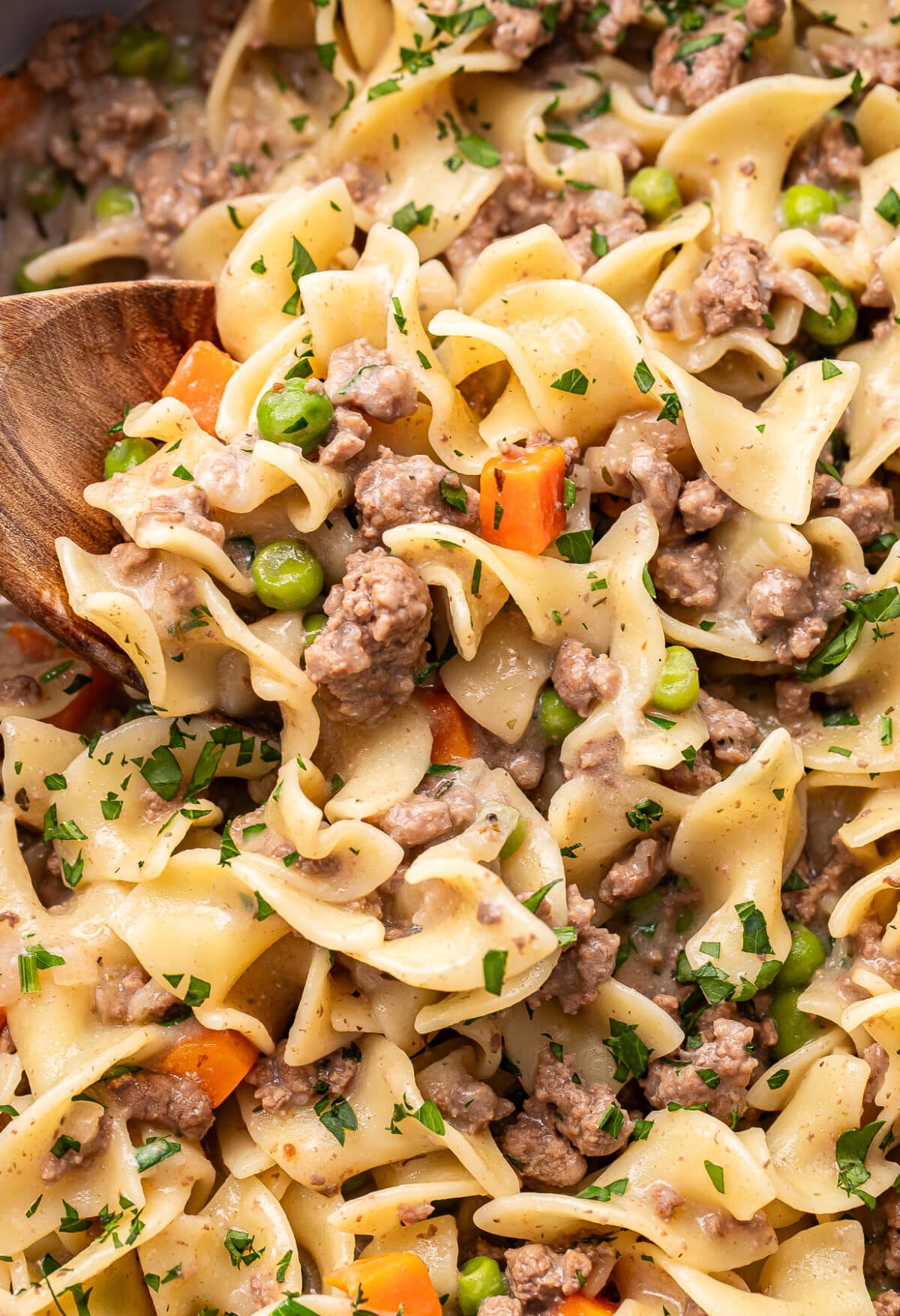 A close-up of beef noodle casserole with ground beef, egg noodles, peas, carrots, and creamy sauce, garnished with chopped parsley. A wooden spoon is mixing the casserole.