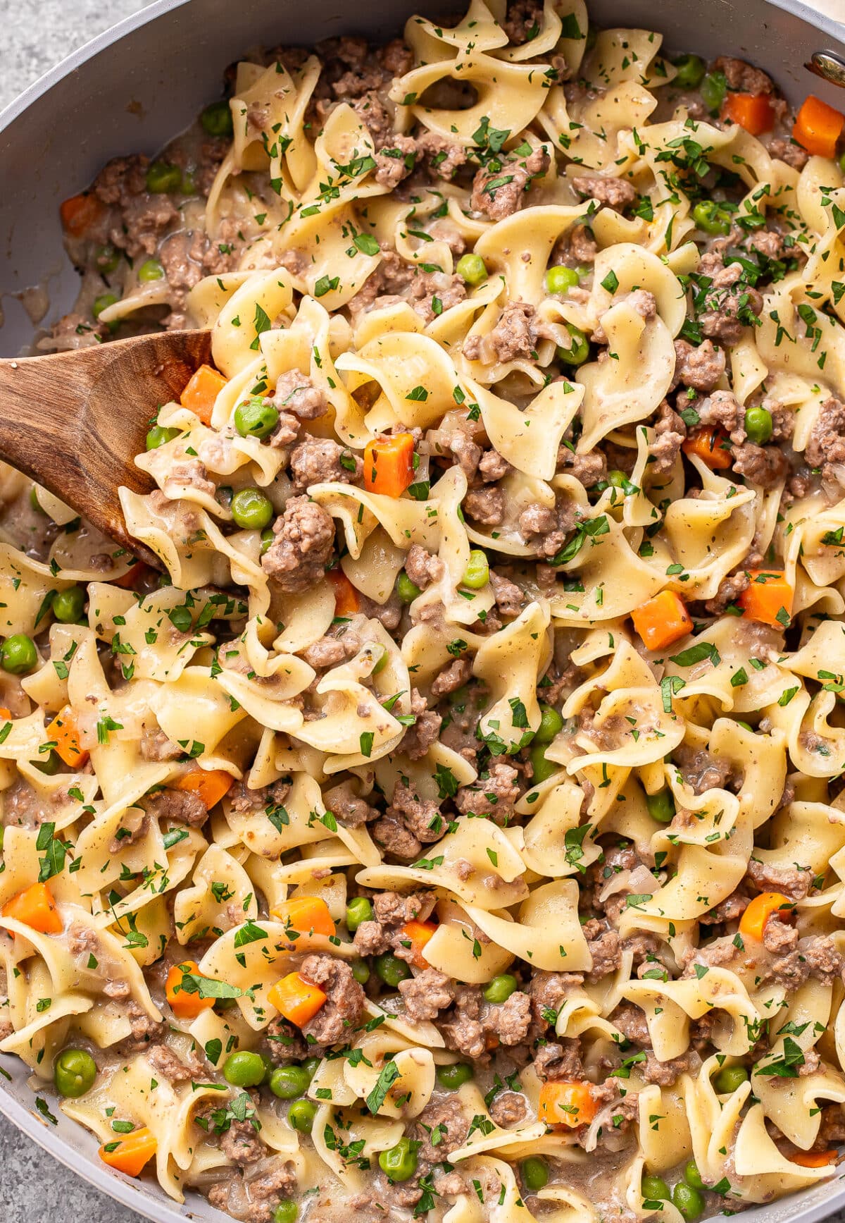 Egg noodles mixed with ground beef, peas, carrots, and a creamy sauce, garnished with chopped parsley, in a pan with a wooden spoon.